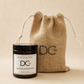 A single wick Pumpkin Marmalade scented candle jar with a black lid and white label next to a natural burlap drawstring pouch, both branded DCw, on a warm beige background.