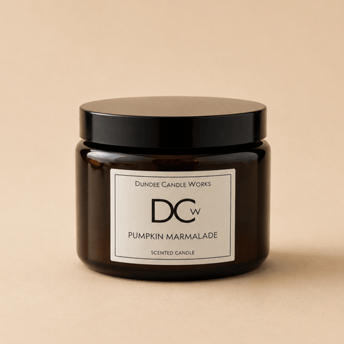 A dark amber glass jar candle with a black lid and a white label reading 'Dundee Candle Works DCw Pumpkin Marmalade Scented Candle' on a warm beige background.