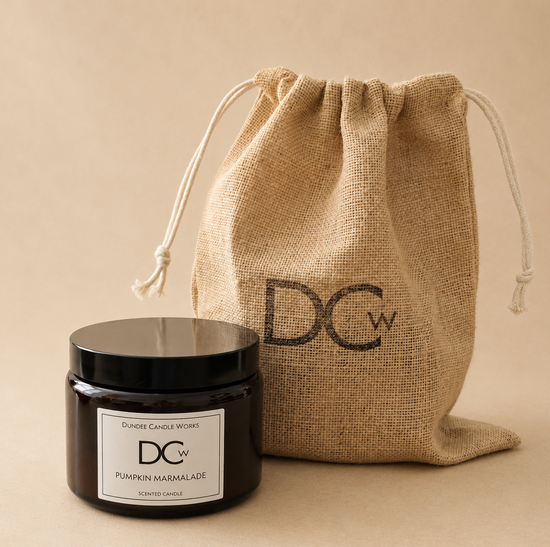 A Dundee Candle Works Pumpkin Marmalade scented candle in a dark glass jar with a black lid and white label sits in front of a natural burlap drawstring pouch printed with the brand logo DCw, on a warm beige background.