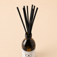 A reed diffuser bottle with a white label and multiple black reeds, photographed against a plain warm beige background. The label shows the brand initials 'DCw' and the name 'Dundee Candle Works'.