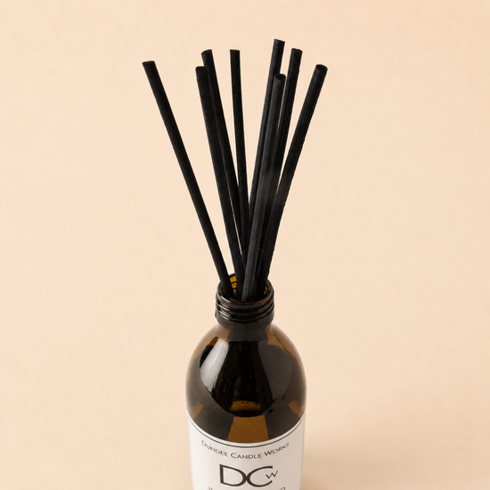 A reed diffuser bottle with a white label and multiple black reeds, photographed against a plain warm beige background. The label shows the brand initials 'DCw' and the name 'Dundee Candle Works'.