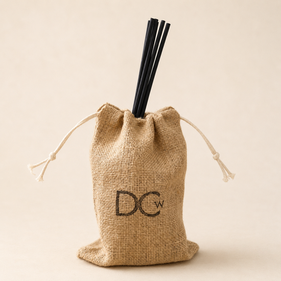 A small natural burlap drawstring pouch with a black monogram logo reading 'DCW' on the front, holding several black reed diffuser sticks, photographed against a warm beige studio background.
