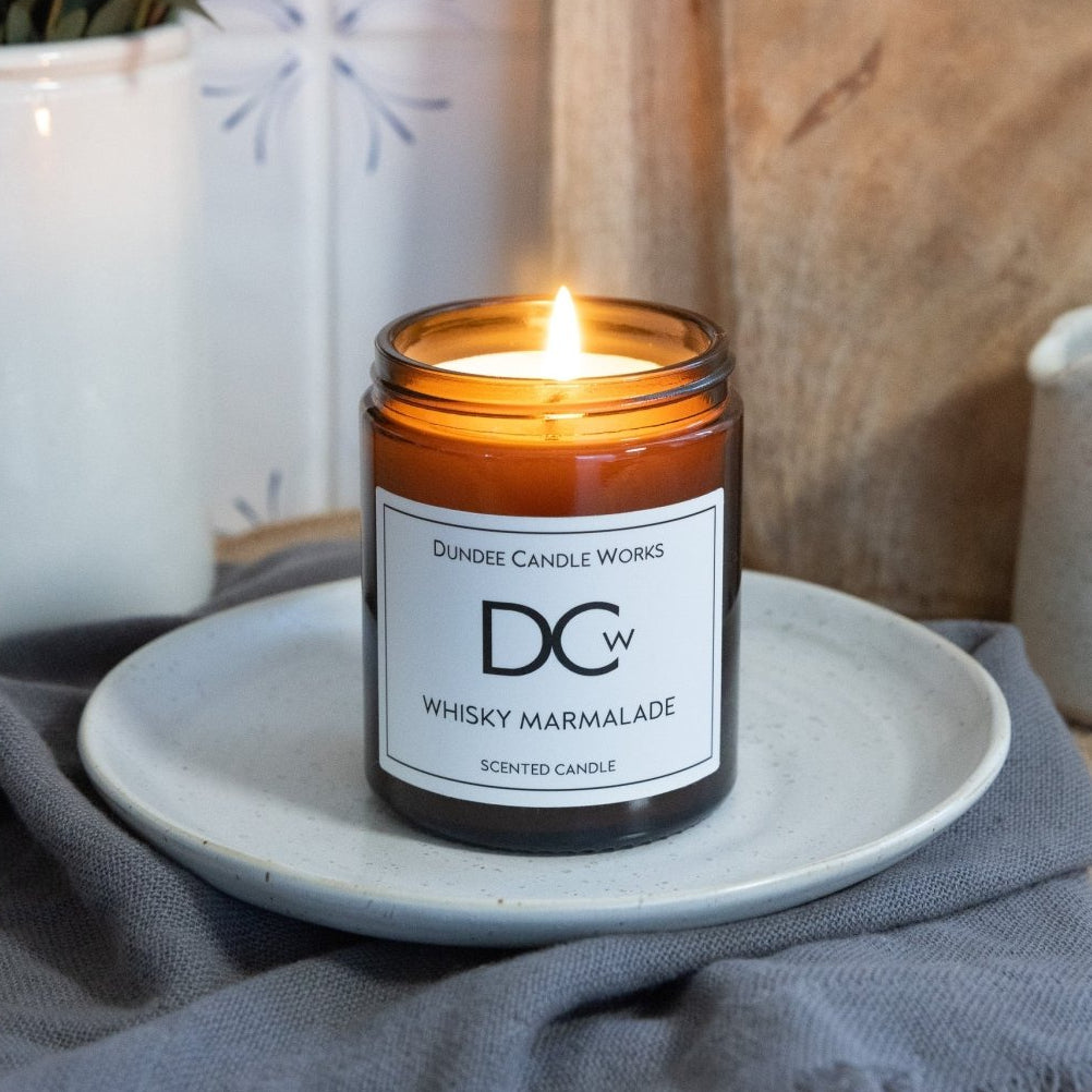 A lit amber glass jar candle labeled 'DCw Dundee Candle Works Whiskey Marmalade' sits on a small white plate on a gray cloth, with a rustic wood background and potted plants nearby.