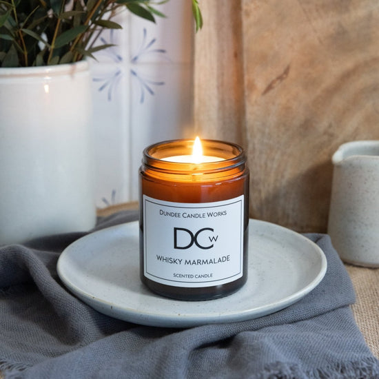 A lit amber glass jar candle labeled 'DCw Dundee Candle Works Whiskey Marmalade' sits on a small white plate on a gray cloth, with a rustic wood background and potted plants nearby.