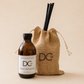 An amber glass bottle labeled Dundee Candle Works DCw Whisky Marmalade reed diffuser, shown next to a natural jute drawstring bag with black reeds and a black DCw logo, on a warm beige background.