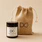 A Whisky Marmalade scented candle in a dark amber glass jar with a black lid and white label, placed in front of a natural burlap drawstring pouch. The pouch and jar both display the DCw logo.
