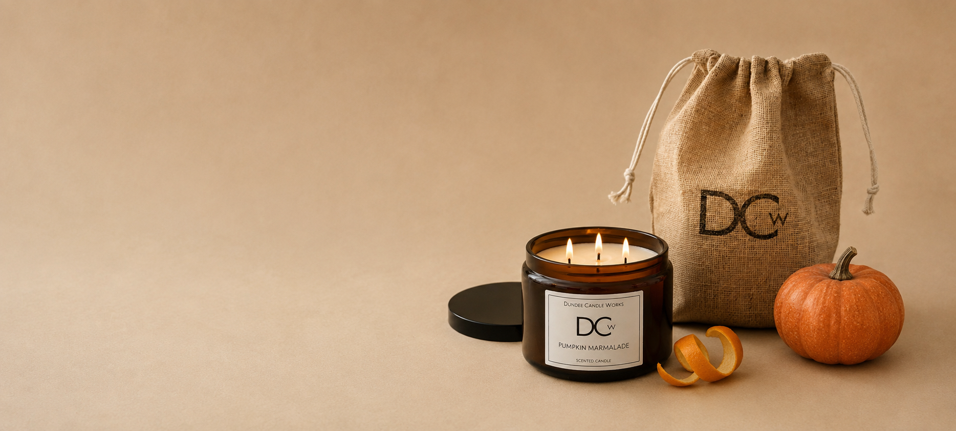 A lit triple-wick amber glass Pumpkin Marmalade candle jar with a black lid beside a burlap drawstring bag and a small orange pumpkin on a warm beige background. The bag and candle label both show the DCw logo.