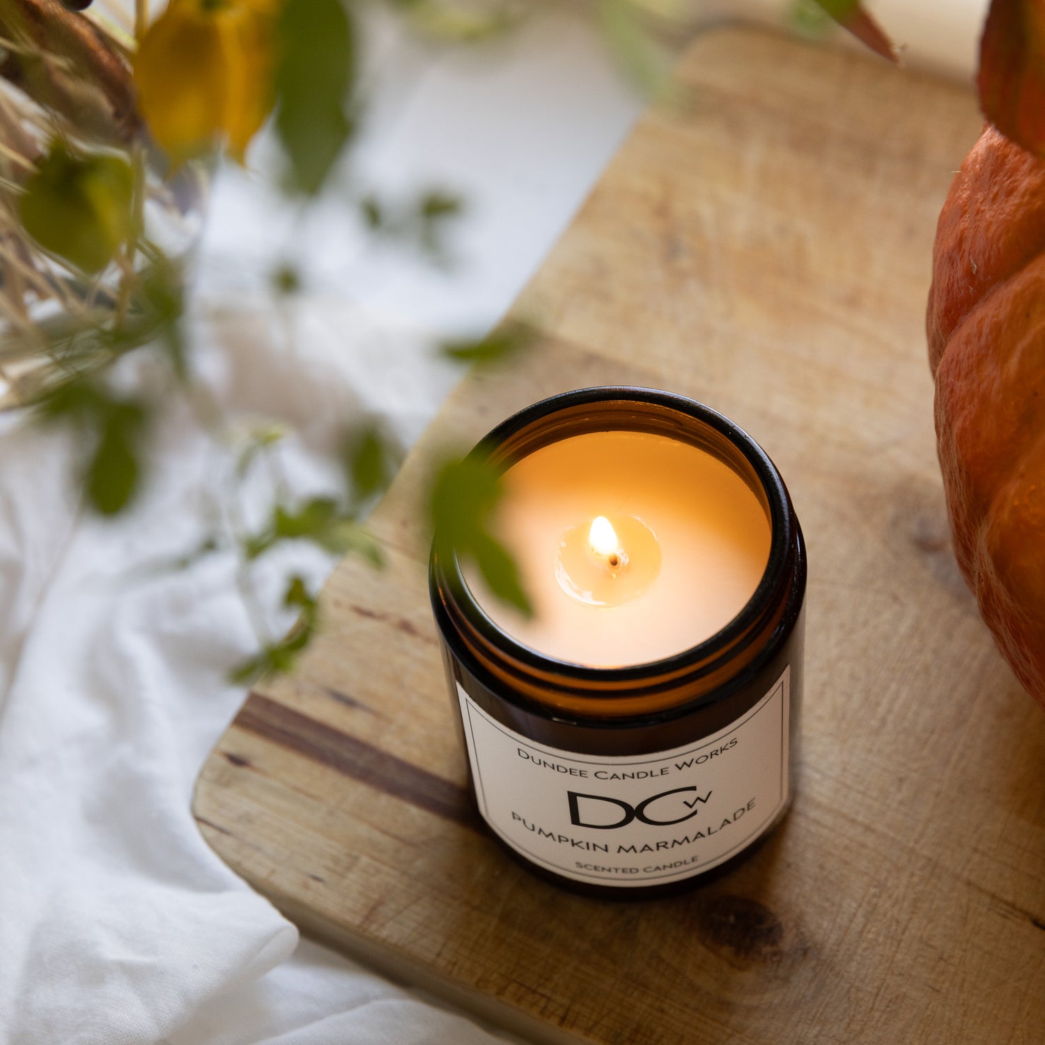 Dundee Candle Works Pumpkin candle in an amber jar on a wooden surface with a pumpkin and plant in the background.
