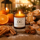 A lit scented candle in a dark amber glass jar sits on a wooden surface surrounded by warm holiday props, including gingerbread cookies, oranges, cinnamon sticks, star anise, and dried orange slices. The front label reads 'Dundee Candle Works' and 'Marmalade Gingerbread'.